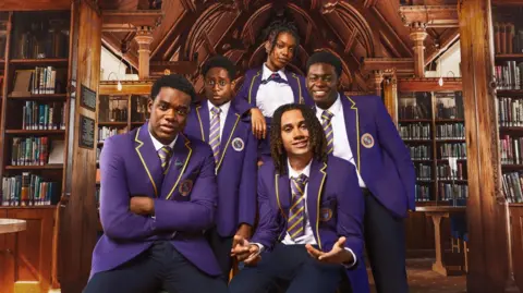 BBC/Studio Lambert The main cast of Boarders, five young people, are standing together. They are all wearing a blue school uniform. They are in an old library with wooden shelves.