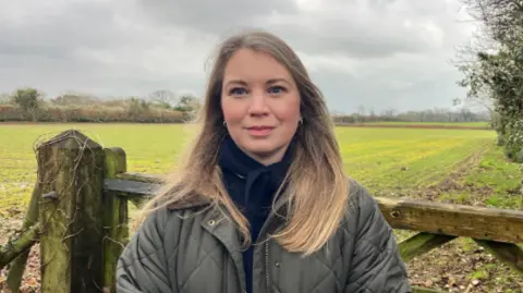 BBC Laura Layzell wearing a green coast with a navy blue top and navy blue scarf. She has long dark blonde hair and is smiling at the camera. Behind her is a field with a wooden gate just before it.