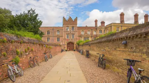 Getty Images A wide shot of the pathway leading to Jesus College at Cambridge University on a sunny day. There are two brick walls on either side of a stone-paved path with pebbles on either side. There are eight bikes symmetrically-placed lined up neatly against the walls