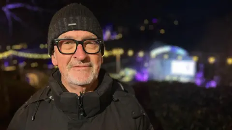 A man with short white facial hair looks at the camera. He wears a black hat, black glasses and a black rain jacket. He is standing outside in the dark in front of a brightly lit stage