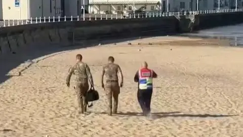 Three people are walking along a sandy beach with seafront buildings in the background. Two are wearing camouflage with one carrying a large black bag, while the third is wearing a high-vis vest.