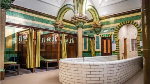 A highly decorated room with green and mustard tiles and ornate columns. There is a deep plunge pool in the foreground with white tiles. At the sides there are booths separated by wooden panels with decorations at the top and green privacy curtains.