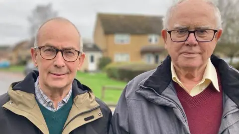 Peter and Chris stand next to each other, looking at the camera. Both wear glasses and waterproof jackets. Peter wears a green jumper and shirt and Chris has a red jumper and shirt.