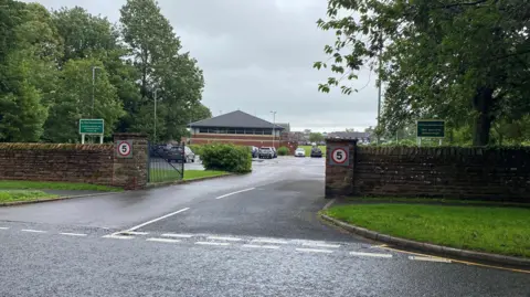 The Nelson Thomlinson secondary school taken from outside the gates shows the entrance with concrete posts on either side. The school's name is on a green sign and in the background the low school buildings which have pyramid roofs.