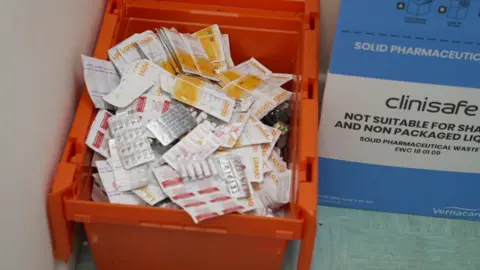 An orange box full of medication on the floor of a pharmacy. A blue and white sign with writing on is next to the box.