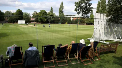 Guests sitting in deckchairs watch a cricket match from behind a black net. A white sight screen is to the right and players can be seen gathered around the batter on a brown cricket strip surrounded by green grass