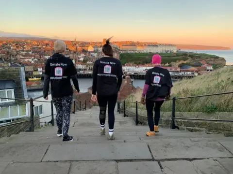 Bryony Jackson, mum Nicky Hurrell and mother-in-law Sally Alexander are in black T-shirts facing down the 199 Whitby Abbey steps and towards the town's harbour