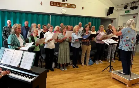 The Voices of Exmoor choir singing in a hall. There is a conductor in the foreground and someone playing a piano.