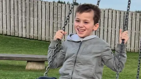 Family photo Max wearing a grey jacket smiles as he sits on a swing.
