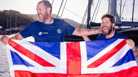 2Stroke.team Karl Austen and Ed Shaw smiling at the camera, wearing navy blue t-shirts and are holding a Great Britain flag.