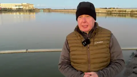Colin Clayton, who chairs the group, is standing in front of the marine lake. He is wearing a black hat, grey jumper and khaki gilet.