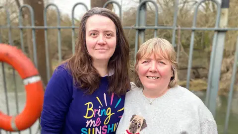 Two women standing side by side. The woman on the left is wearing a top reading "Bring Me Sunshine"; the woman on the right is wearing a jumper with a picture of a pug dog. There is a lifesaving ring and railings in the background.