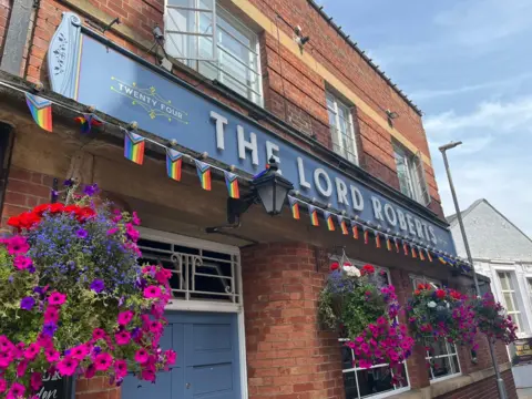 A pub with a sign which says 'the lord roberts'. Underneath the sign there are brightly coloured flowers and small Pride flags. 