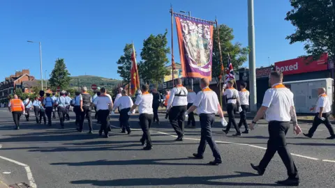 An Orange Order parade passing the Ardoyne shop fronts in north Belfast. There are a number of men in white shirt and black trousers wearing their Orange lodge sashes.  We can only see their backs. There is a large banner in the middle of the group being carried by two men on either side of it. You can see some hills off in the distance.