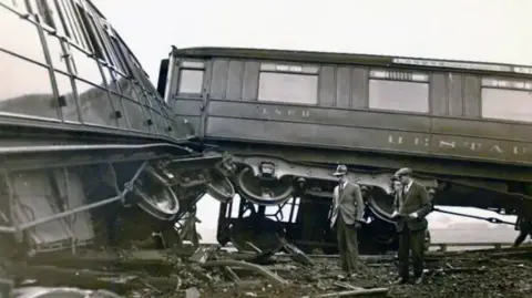 Working Class Movement Library A black and white image of a train crash from 1928 with two carriages off the line. Two men wearing bowler hats stand in the wreckage