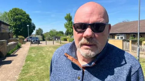 A bald man with dark glasses and a goatee wearing a blue jumper looks at the camera.
