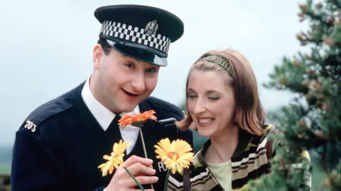 Andrew Agnew, as PC Plum, and Julie Wilson Nimmo, as Miss Hoolie the nursery teacher, smell some flowers