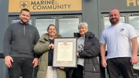 Two women are standing next to each other and holding a framed certificate, while two men stand to either side of them. A building with a sign saying "Marine in the community" is behind them.