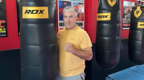 TOM LARSEN-WRIGHT/BBC Steve Kerridge poses next to a punching bag with his fist raised. He is wearing a yellow T-shirt and looking directly at the camera. 