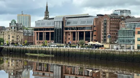Newcastle Crown Court reflected in the River Tyne running in front of it. It is an imposing building made from smooth red stone with massive black windows and tall columns along its frontage.