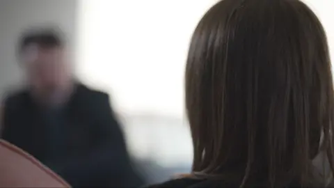 The back of a woman's head in the foreground. She has brunette hair. Out of focus in the background is reporter.