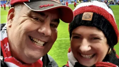 A family photograph showing Ian Wake, on the left, wearing a red and black SAFC branded cap, and a red and white scarf around his neck. On the right, his friend Michelle, is wearing a black, red and white wool hat and a similar red and white coloured scarf. They are both standing on a football pitch. 