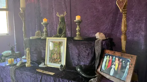 An altar covered with purple velvet cloth. A goddess statue with antlers sits in the middle with other images and statues of goddesses. There are big candles, crystals and incense.