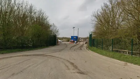 Google A road is leading to a landfill site. There are trees either side of the road which are behind a green fence. A small blue building is in the middle of the road.