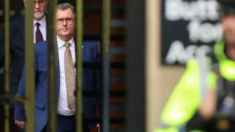 Jeffrey Dolandson in a blue suit and glasses seen through bars aof an entrance gate with a police man in neon yellow in the foreground