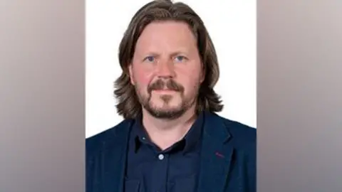 OMBC Mark Kenyon poses for a council portrait. He has shoulder length brown hair and a beard, and is wearing a blue blazer and blue shirt. 