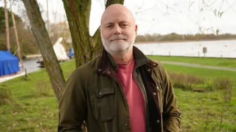 Shaun Whitmore/BBC John Currie is wearing a green wax jacket and a light red T-shirt. He has a grey beard and is looking directly at the camera, standing on the banks of the Broads.