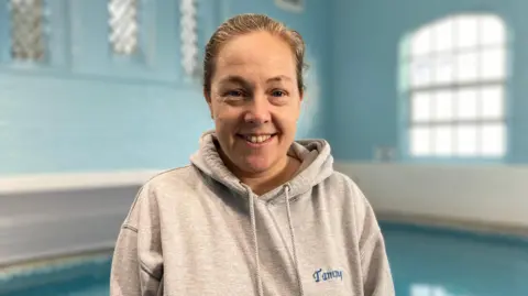 Tammy Harrison, wearing a grey branded hoodie, smiles at the camera with the pool in the background. 