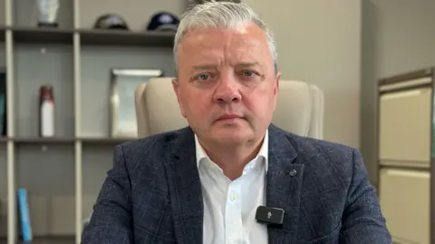 BBC Darragh Cullen, a man with short, white hair, looks tot he camera with a serious expression. He is wearing a navy checked suit jacket and a white shirt. He is sitting in an office.