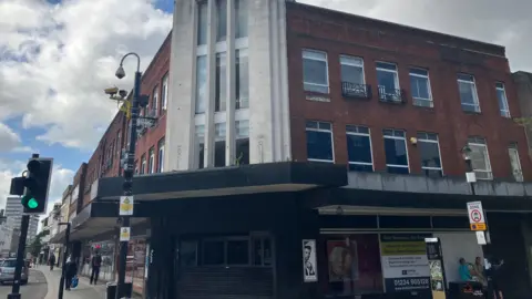 Amy Holmes/BBC A corner building with an Art Deco–style tower featuring tall vertical windows. The rest of the structure is red brick with rows of rectangular windows. Ground‑floor shopfronts and street signs line the pavement, with a traffic light and a few people visible nearby under a cloudy sky.