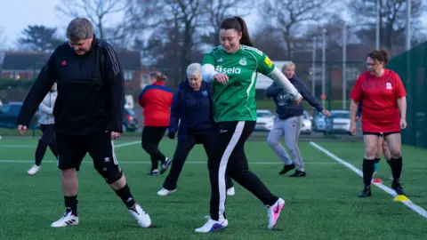 Simon Thurlow To the left is a woman with short hair, dark sports top, dark shorts, dark socks and white football boots, with Sarah Lewis to her left who has long dark hair, green sports top, black and white sports bottoms and white football boots. There are five other women on football pitch behind them. They are all doing training exercises.