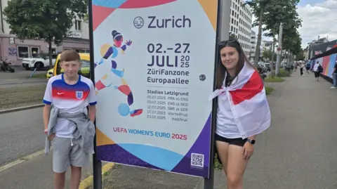 Family Ethan and Amelia stand next to a sign advertising dates for the games. Amelia is draped in an England flag.