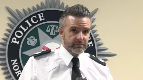 Deputy Chief Constable Bobby Singleton. He has short black and grey hair and is wearing a police uniform. He is standing in front of the PSNI crest.