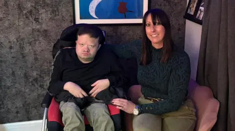 Family handout A young man sits in a wheelchair looking at the camera. A woman is sitting next to him on an armchair, with her arm around him. She is smiling at the camera.