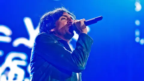 BBC Snow Patrol singer Gary Lightbody sings into a microphone on stage wearing a blue jacket. Behind him is a blue screen with white letters.