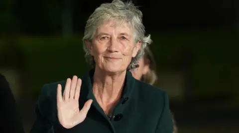 PA Media A woman with short grey hair waves to the camera. She is wearing a dark green coat. The background is dark and out of focus. 