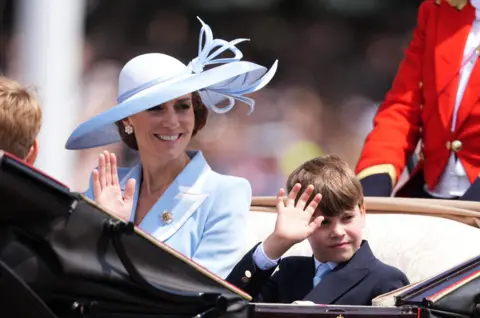 PA Media Princess Catherine smiles while sitting next to Prince Louis in a carriage, She is wearing a light blue wide-brimmed hat and a light blue jacket with white-trimmed lapels and a brooch. Louis is wearing a suit with a light blue tie.
