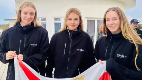 Jersey Sport Three girls, all with long light brown hair, and wearing black fleeces, holding a Jersey flag.