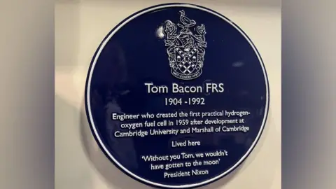 Emma Baugh/BBC A blue plaque on a cream wall. It recognises the achievements made by scientist Tom Bacon and includes a quote from President Nixon