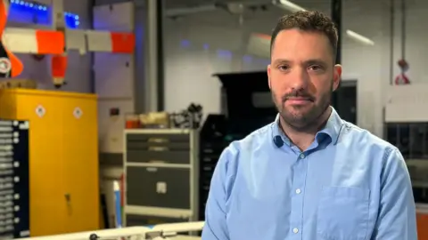 Dr Chris Holmes is wearing a blue button up shirt, and he has short brown hair. Behind him is an out of focus workshop.