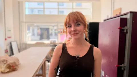 A young women with red hair done up in a bun. she is wearing a brown tank top and a triangle pendant necklace. she is looking at the camera smiling.