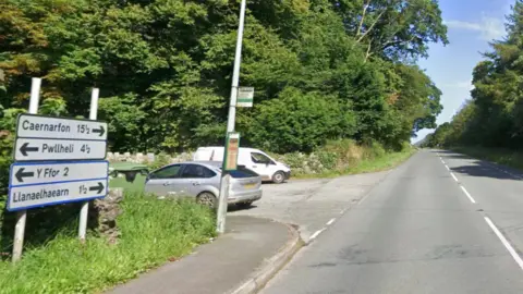A499 showing road signs pointing to Pwllehli in one direction and Y Ffor other way. Parked cars and a bus stop seen on the left.