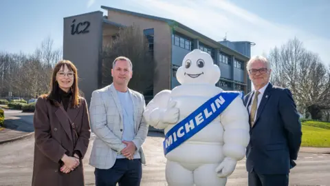 A woman and two men are standing in front of a large building alongside a large white padded figure with the word "Michelin" on a sash in front of it.