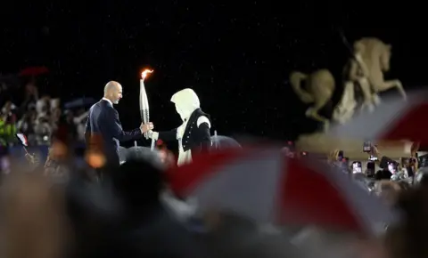   Cameron Spencer/Getty Images The torchbearer hands over the torch to former French footballer Zinedine Zidane at the Place du Trocadero during the opening ceremony of the Olympic Games