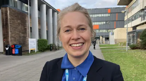 Dr Munro is standing outside Aberystwyth University, with buildings and grass behind her. She is wearing a blue blouse and black blazer and is smiling. It is a head and shoulders shot.