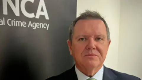 Gerry McLean standing in front of a National Crime Agency sign, wearing a dark suit, white shirt and a green tie.